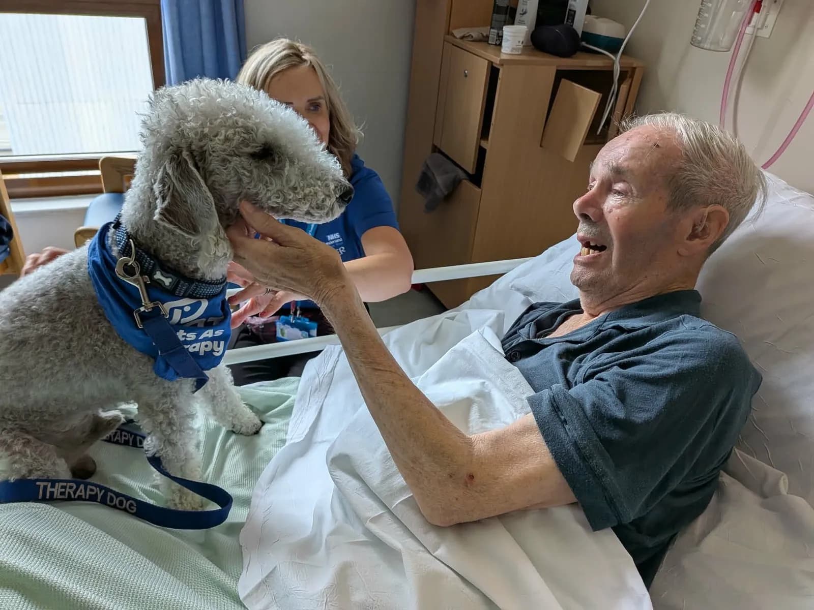 A man in a hospital bed smiles and reaches out to pet a therapy dog with a blue bandana and leash, while a woman in a blue shirt stands behind the dog.